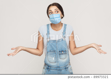 I don't know. Portrait of thoughtful young brunette woman with surgical medical mask in denim overalls standing raised arms and looking away. indoor studio shot isolated on gray background. I don't know. Portrait of thoughtful young brunette woman with surgical medical mask in denim overalls standing raised arms and looking away. indoor studio shot isolated on gray background. 89973321
