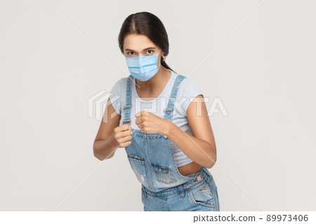 Portrait of angry mad young brunette woman with medical mask in denim overalls standing with boxing fists, looking and ready to attack or defence. indoor studio shot isolated on gray background. 89973406