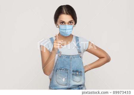 Portrait of serious young brunette woman with surgical medical mask in denim overalls standing looking at camera and warning or scolding. indoor studio shot isolated on gray background. Portrait of serious young brunette woman with surgical medical mask in denim overalls standing looking at camera and warning or scolding. indoor studio shot isolated on gray background. 89973433