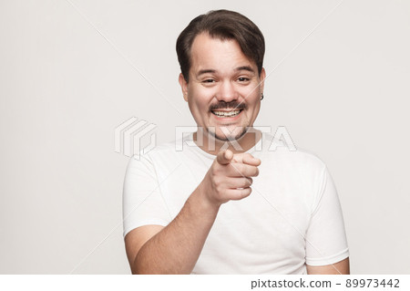 The happiness man, wearing white tshirt, having happiness looks, pointing finger at camera and toothy smiling. Isolated studio shot on gray background 89973442