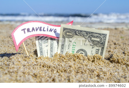 Paper bills one dollar and small stick with paper speech bubble with word Traveling buried in sand beach background sea close-up sunny summer day. Concept Money travel tourism vacation holiday relax. Paper bills one dollar and small stick with paper speech bubble with word Traveling buried in sand beach background sea close-up sunny summer day. Concept Money travel tourism vacation holiday relax. 89974602