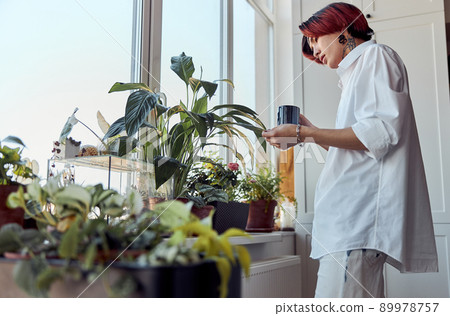 Asian guy holding cup of coffee and touching the plants on the windowsill 89978757