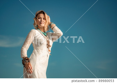 Woman in bohemian clothing on beach at sunset. Boho style for fashionable look on resort. Middle aged well looking woman in white dress and boho style braclets. Summer fashion Woman in bohemian clothing on beach at sunset. Boho style for fashionable look on resort. Middle aged well looking woman in white dress and boho style braclets. Summer fashion 89980053