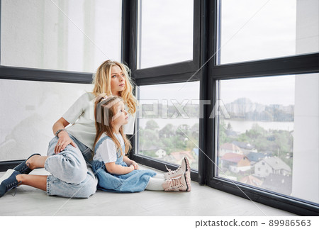 Woman and little girl sitting on the floor and looking out window in family new home. Mother and child enjoying view from panoramic window in newly built apartment. 89986563