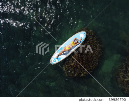 Top view on blonde woman in yellow bikini relaxing on the sup board. Young attractive woman in the sea on the Stand Up Paddle Board SUP. The concept of active and healthy life in harmony with nature. Top view on blonde woman in yellow bikini relaxing on the sup board. Young attractive woman in the sea on the Stand Up Paddle Board SUP. The concept of active and healthy life in harmony with nature. 89986790
