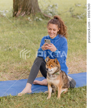 woman with a dog in the park does sports or fitness on a yoga mat. a European woman with curly hair in a blue hoodie is chatting on a smartphone 89987690