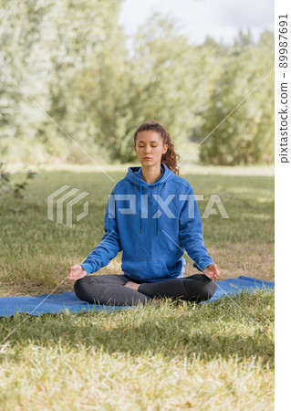 woman in the park does yoga, sports, meditation or fitness on a yoga mat. a European woman with curly hair in a blue hoodie takes care of her body and mental health woman in the park does yoga, sports, meditation or fitness on a yoga mat. a European woman with curly hair in a blue hoodie takes care of her body and mental health 89987691