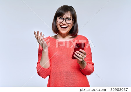 Portrait of middle aged beautiful woman with phone in hands looking at camera on light background Portrait of middle aged beautiful woman with phone in hands looking at camera on light background 89988087