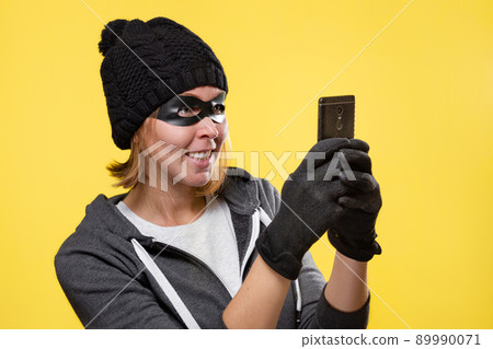 The concept of cybercrime and hacking. Portrait of a woman in a black hat, gloves and mask, who holds a mobile phone and smiles ominously. Yellow background. Copy space The concept of cybercrime and hacking. Portrait of a woman in a black hat, gloves and mask, who holds a mobile phone and smiles ominously. Yellow background. Copy space 89990071
