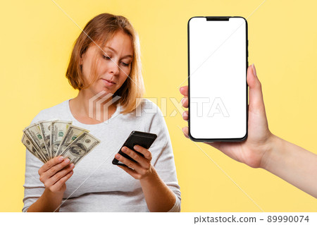 A young woman holds a fan of dollars and a cellphone. On the right is a smartphone with a white screen. Mock up. Yellow background. Online Shopping concept 89990074