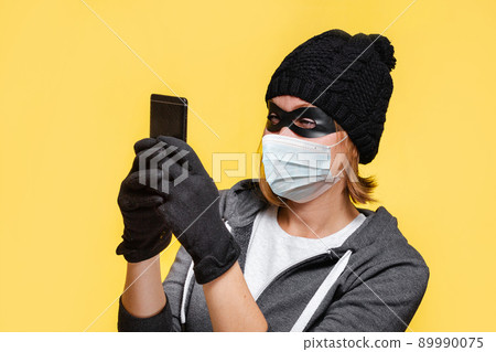 A female hacker in a black hat, gloves and a protective mask holds a smartphone in her hands, trying to hack it. Yellow background. The concept of cybercrime and new normal A female hacker in a black hat, gloves and a protective mask holds a smartphone in her hands, trying to hack it. Yellow background. The concept of cybercrime and new normal 89990075