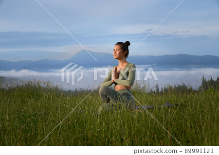 Sporty young woman sitting on yoga mat with namaste gesture and enjoying outdoors meditation. Concept of wellness, spirituality and harmony. Sporty young woman sitting on yoga mat with namaste gesture and enjoying outdoors meditation. Concept of wellness, spirituality and harmony. 89991121