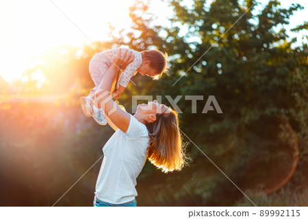 Young happy mother throws up baby in the sky, summer outdoors. Side view. Concept of happy childhood 89992115