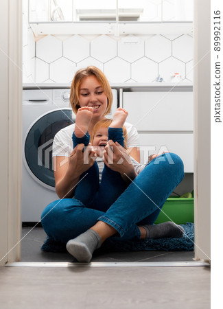 Happy mother and her little daughter play together in the bathroom, sitting on the floor. Vertical. Washing machine in the background 89992116