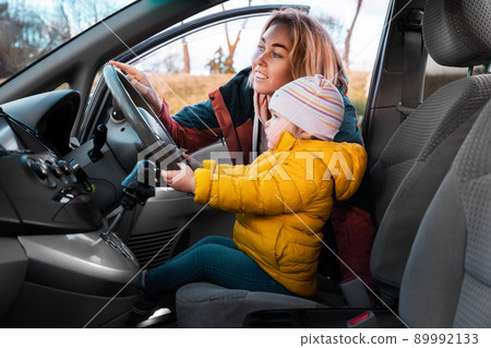 A young smiling mother teaches her little child to drive a car. The kid is sitting in a chair and holding the steering wheel with his hands. Side view 89992133