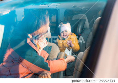 Mother in a protective mask, sitting in car and giving a mask to her child sitting in the back seat in a child car seat. View through the windshield 89992134
