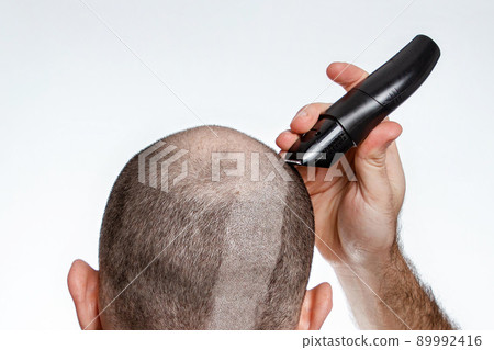 The concept of baldness and alopecia. A man holds a hair clipper and shaves the hair on top of his head. The view from the back. Close up The concept of baldness and alopecia. A man holds a hair clipper and shaves the hair on top of his head. The view from the back. Close up 89992416