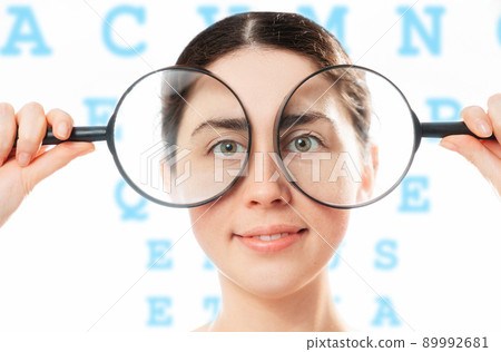 Ophthalmology. Portrait of a young smiling pretty woman holding magnifying glasses to her eyes. The alphabet is in the background. The concept of healthy eyes and an optical test 89992681