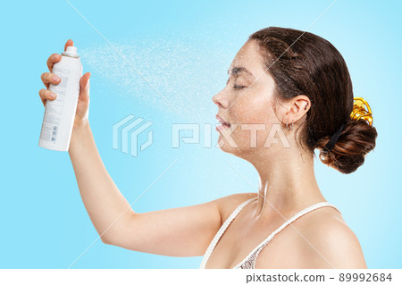 A young Caucasian woman with wet skin sprays thermal water on her face. Blue background. Side view. The concept of cosmetology, skin care and moisturizing A young Caucasian woman with wet skin sprays thermal water on her face. Blue background. Side view. The concept of cosmetology, skin care and moisturizing 89992684