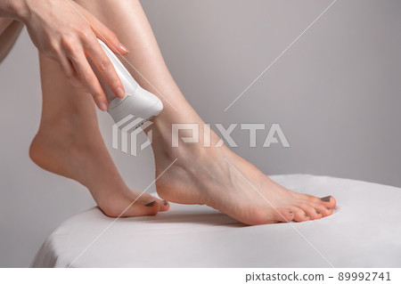 A young woman removes excess hair with a depilator. Concept of home skin care and Spa. Smooth slender legs. White and grey background. Close up and copy space 89992741