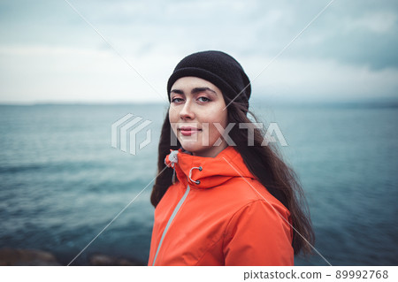 Portrait of a smiling brunette woman with long hair, wearing a hat and an orange jacket. In the background the sea and the horizon line. Copy space 89992768