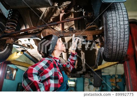 Portrait of a young woman mechanic conducting an inspection of the car body. Side view. Bottom view 89992775