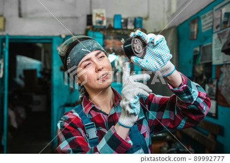 Portrait of a young female mechanic in uniform, wearing gloves and safety glasses on her head, checking a car part. Garage in the background 89992777