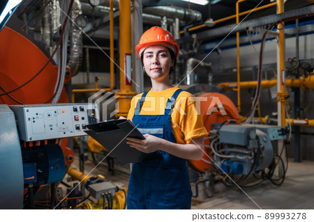 Concept of a industrial manufactory. A female smiling worker in uniform and helmet with a folder and a pen in her hand. In the background-boiler room 89993278
