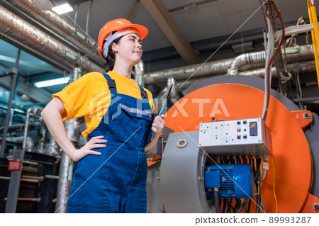A young worker in a helmet and uniform holds a folder in her hand and stands in the boiler room. Mock up. Bottom view. The concept of industrial production A young worker in a helmet and uniform holds a folder in her hand and stands in the boiler room. Mock up. Bottom view. The concept of industrial production 89993287