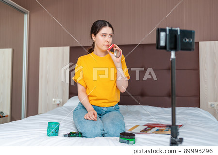 Vlogging. A beautiful female vlogger streaming live video on smartphone sitting on bed and smells a new cosmetic. Home related 89993296