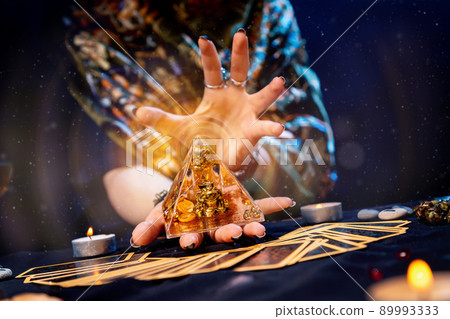 The fortune teller holds a glass pyramid with gold in her hands and casts a spell over it. Magic aura. There are Tarot cards on the table. Close-up. The concept of divination, magic and esotericism 89993333