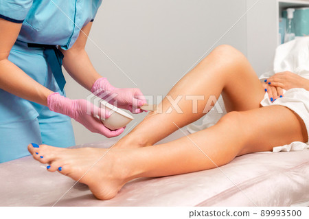 Epilation concept. Cosmetologist in pink medical gloves spread agent gel to the client's feet. Hair removal in a professional beauty salon 89993500