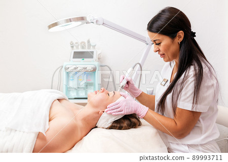 Beauty salon. A cosmetologist in rubber gloves does a aqua peeling procedure on the face. In the background, a cosmetology device. Concept of professional cosmetology and beauty Beauty salon. A cosmetologist in rubber gloves does a aqua peeling procedure on the face. In the background, a cosmetology device. Concept of professional cosmetology and beauty 89993711