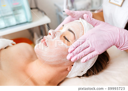 Cosmetologist's hands at protective gloves massages client's face with cosmetics mask. Concept of professional cosmetology 89993723