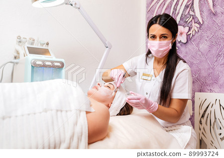 New normal. Professional female cosmetologist in a protective mask and gloves, applies the mask to the client's face. The concept of professional cosmetology service New normal. Professional female cosmetologist in a protective mask and gloves, applies the mask to the client's face. The concept of professional cosmetology service 89993724