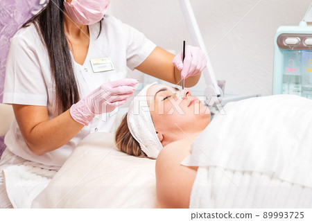 A cosmetologist in a medical mask and gloves holds a bowl of cosmetics, applying a brush mask on the client's face. Side view. Copy space. Concept of professional cosmetology during coronavirus 89993725