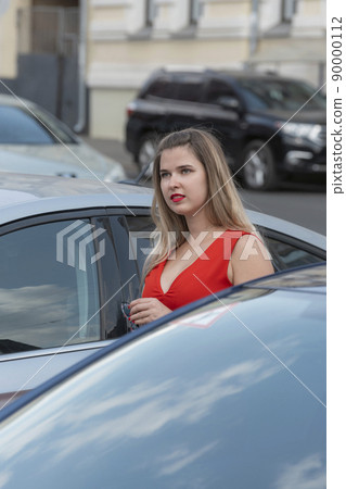 Young attractive girl wears red dress with deep neckline and red lipstick near cars. Vertical frame. 90000112