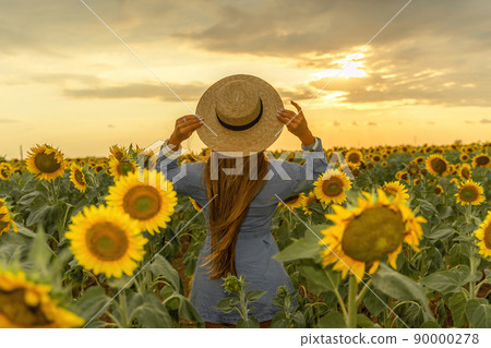Beautiful middle aged woman looks good in a hat enjoying nature in a field of sunflowers at sunset. Summer. Attractive brunette with long healthy hair. 90000278