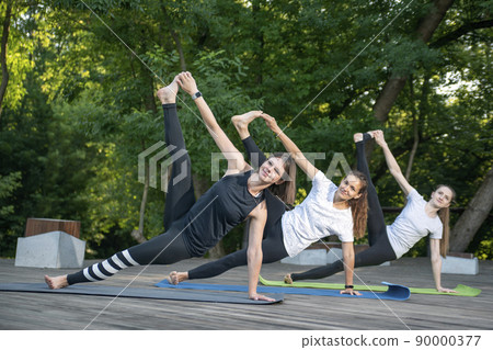 Three young woman workout in summer park doing exercise outdoor. Women does yoga asana together outside. 90000377