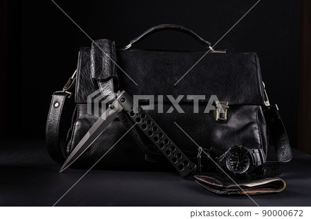 A large black folding knife on the background of a black leather bag. Gentleman's set of knife, watch and wallet. Knife in half-folded position. A large black folding knife on the background of a black leather bag. Gentleman's set of knife, watch and wallet. Knife in half-folded position. 90000672
