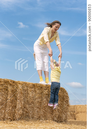 Young mother plays with small child on farm in dry hay. Mom and daughter on haystack. Blue sky. Summer day. Young mother plays with small child on farm in dry hay. Mom and daughter on haystack. Blue sky. Summer day. 90000853