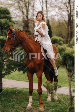 A woman in a white sundress riding a horse near a farm A woman in a white sundress riding a horse near a farm 90003370
