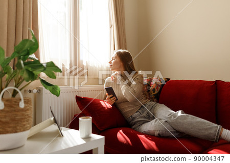 Young woman using smartphone at cozy home on sofa in living room, side view, sunlight from window. Young woman using smartphone at cozy home on sofa in living room, side view, sunlight from window. 90004347