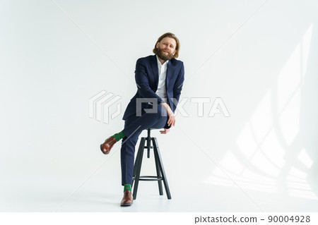 A handsome brutal bearded and long-haired man, a stylish businessman sitting on a chair with a white background 90004928