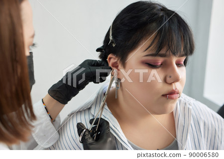 A beautician makes a puncture of the earlobe with a disposable device in a beauty salon. Ear piercing lobe. 90006580