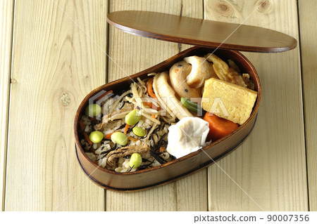 A bird's-eye view of the Makunouchi lunch box containing a lot of side dishes such as hijiki and edamame rice, shumai, omelet, and simmered dishes. 90007356