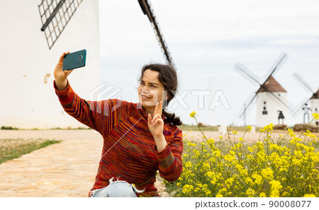 Cheerful woman traveler with smartphone near windmills in Mota del Cuervo 90008077