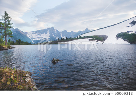 Mountain valley with snow forest and lake at foot. Rocks are reflected in river. Melting glacier on slopes of ridge. Dawn clouds foreshadow precipitation. 90009318