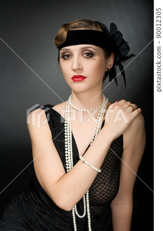 Portrait of a glamorous dark haired model with perfect makeup wearing a luxurious necklace, black bodysuit and long top, holding her hands behind her head and looking straight ahead. 90012305