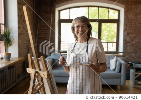 Older painter woman posing near easel with palette and paintbrush 90014421